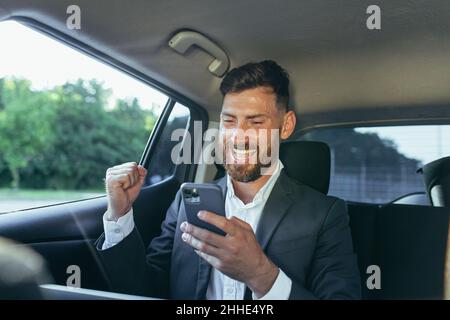 Homme d'affaires heureux et réussi passager de voiture lit les nouvelles du téléphone, se réjouit de la victoire et les nouvelles reçues Banque D'Images