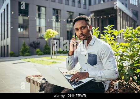 de jeunes étudiants africains étudient en ligne sur un banc près de l'université, utilisent un ordinateur portable et un smartphone Banque D'Images