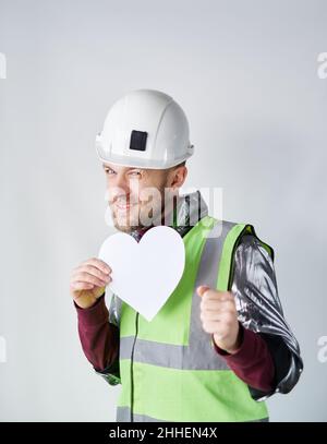 Saint-Valentin, romance ou amour concept.Dansant barbu homme travailleur, électricien ou technicien en casque de sécurité et gilet réfléchissant tenant le papier blanc coeur forme.Image verticale de haute qualité Banque D'Images