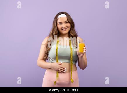 Bonne jolie caucasienne plus grande taille jeune femme dans les vêtements de sport avec mètre ruban montrer le verre de jus d'orange frais