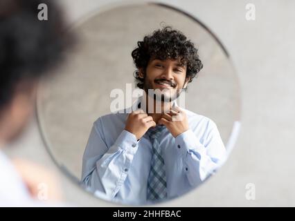 Un homme d'affaires indien enthousiaste se prépare et se fesse une chemise, debout dans la salle de bains devant le miroir Banque D'Images