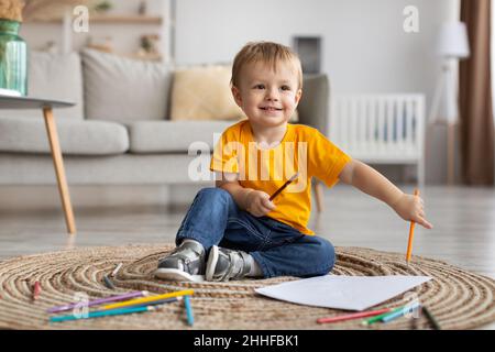 Portrait d'un jeune garçon heureux assis sur un tapis avec des crayons colorés et une feuille de papier, dessin à la maison, espace de copie Banque D'Images