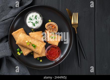 Fines crêpes farcies à la viande et aux carottes aux herbes fraîches, à la crème aigre et au caviar rouge dans une assiette noire sur fond de bois. Banque D'Images