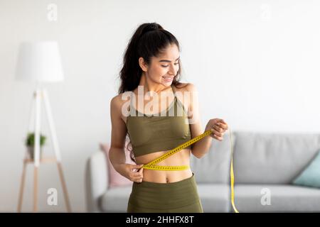 Concept de perte de poids réussi.S'adapte à la jeune femme indienne dans des vêtements de sport mesurant la taille avec du ruban adhésif à la maison Banque D'Images