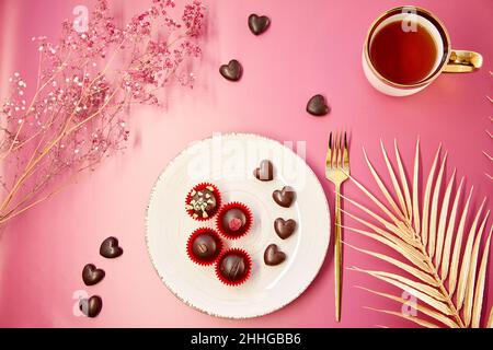 Cœurs de chocolat et bonbons décorés sur l'assiette ancienne blanche avec fourchette et tasse de thé.Dîner romantique, concept délicat de la Saint Valentin.Fond rose, gitsophila et décorations dorées. Banque D'Images