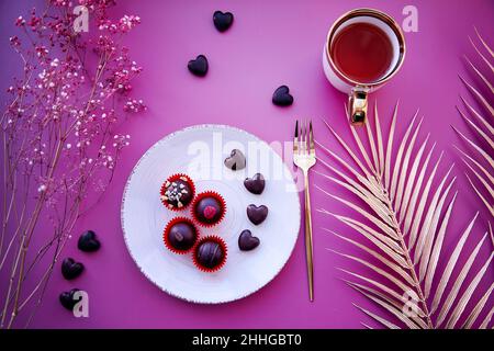 Cœurs de chocolat et bonbons décorés sur l'assiette ancienne blanche avec fourchette et tasse de thé.Dîner romantique, concept de la Saint-Valentin.Fond violet, gitsophila et décorations dorées.Vue de dessus Banque D'Images