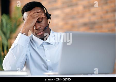 Gros plan d'un homme afro-américain fatigué avec un casque, un expert, un consultant, un opérateur de ligne directe, tenant sa tête,trop fatiguée d'un appel long et lourd.Concept de service d'assistance, assistance téléphonique, communication à distance. Banque D'Images