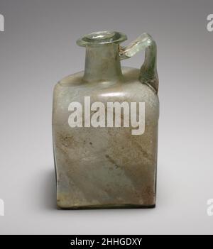 Flacon carré en verre fin 1st–3rd Century A.D. Roman translucide vert pâle, avec manche de même couleur.rebord replié, au-dessus et à l'intérieur, formant légèrement une constriction à la bouche, avec surface supérieure biseautée; col cylindrique, avec repères d'outillage autour de la base; épaulement enfoncé avec coins arrondis et inclinés; côtés plats et verticaux; fond plat;Large poignée à lanière, avec décoration peignée sur la surface extérieure, fixée au bord extérieur de l'épaule, dressée verticalement, tournée en angle aigu et tractée sur le cou, avec une traînée se terminant sur le dessous de la jante. Décoration moulée sur le fond comprenant quatre angles en forme de L au niveau du maïs Banque D'Images