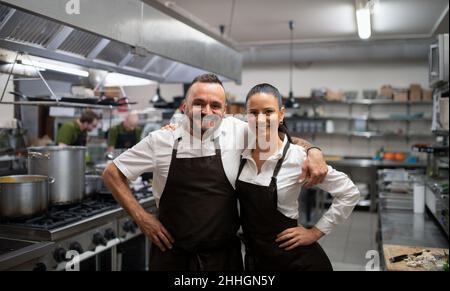 Des chefs confient des collègues qui se tiennent debout avec les bras et regardent l'appareil photo dans une cuisine commerciale. Banque D'Images