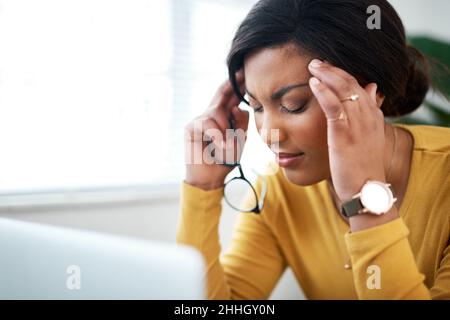 Ai-je trop fait.Photo courte d'une jeune femme attrayante assise à la maison et souffrant de maux de tête tout en utilisant son ordinateur portable. Banque D'Images
