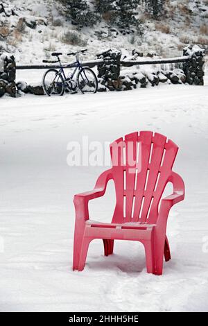 Une chaise de pelouse rouge a été laissée dans la cour pendant une tempête de neige, avec un ancien VTT appuyé contre une clôture en bois en arrière-plan. Banque D'Images