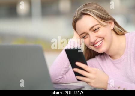 Heureux adolescent utilisant un smartphone et un ordinateur portable assis dans la rue Banque D'Images