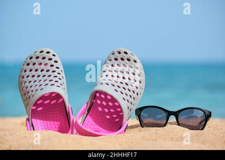 Gros plan sur les chaussures Clogs et les lunettes de soleil noires protectrices sur la plage de sable au bord de la mer tropicale par temps chaud et ensoleillé. Concept de vacances d'été. Banque D'Images