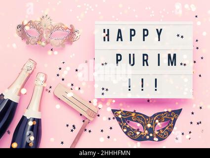 Concept de célébration du Carnaval de Purim.Happy Purim écrit en boîte lumineuse, bouteilles de champagne, masque de carnaval et gragger en bois sur fond rose Banque D'Images