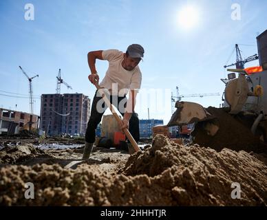 Workman travaille sur le chantier de construction de rue et utilise une pelle pour remplir la machine à pétrir avec un mélange ciment-sable.Processus de préparation du matériau pour la table semi-sèche. Banque D'Images