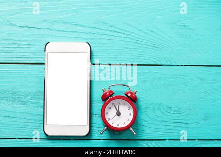 Téléphone mobile et réveil rétro rouge avec cinq minutes à douze heures sur fond bleu en bois.Vue de dessus et écran vide Banque D'Images