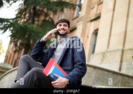 D'en-dessous, un homme heureux élève barbu avec des livres assis sur l'escalier en pierre du bâtiment de l'université et le téléphone cellulaire parlant Banque D'Images