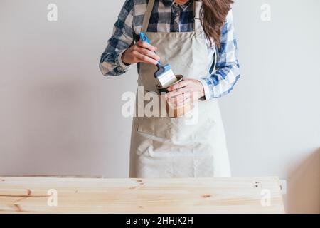 Croisez une menuiserie féminine non reconnaissable dans un tablier avec un pot de fini bois et brossez-vous près d'une planche en bois sur fond blanc Banque D'Images