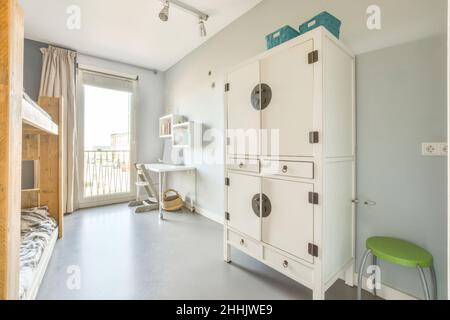 Intérieur de la chambre lumineuse pour enfants avec lits superposés en bois face de l'armoire et bureau blanc avec étagères au mur près de la fenêtre pleine longueur Banque D'Images