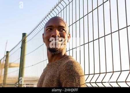 Un homme afro-américain positif regarde loin en se tenant près d'une clôture métallique sur une rue ensoleillée contre un ciel sans nuages dans la ville Banque D'Images