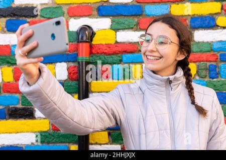 Une jeune touriste ravie avec une longue tresse en veste chaude et des lunettes de vue tout en emportant un selfie sur un téléphone portable près de la dura murale multicolore Banque D'Images