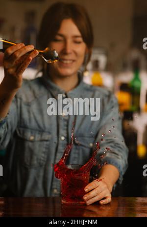 Femme barman ajoutant des glaçons dans le verre avec un cocktail alcoolisé à l'ancienne tout en se tenant au comptoir du bar pendant le travail Banque D'Images