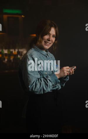 Bonne serveuse regardant l'appareil photo et prenant la commande en écrivant dans le carnet près du comptoir du bar pendant le travail dans un café moderne Banque D'Images