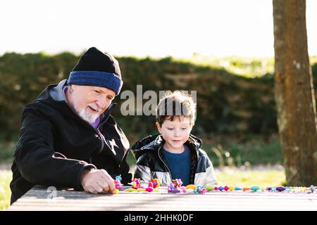 Grand-père barbu en vêtements d'extérieur assis à la table tout en jouant des jouets avec petit-fils dans le parc Banque D'Images