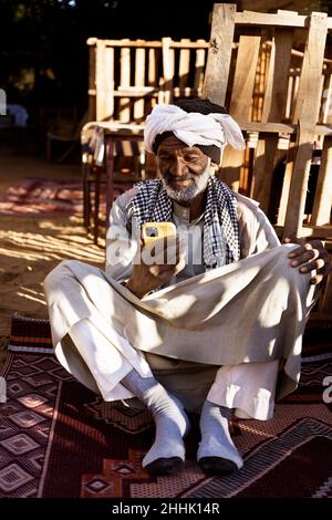 Homme nubien âgé en vêtements traditionnels et en turban assis dans la cour et utilisant le téléphone portable le jour ensoleillé Banque D'Images