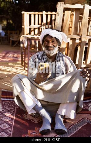 Homme nubien âgé dans des vêtements traditionnels et turban assis regardant la caméra dans la cour et utilisant le téléphone portable le jour ensoleillé Banque D'Images