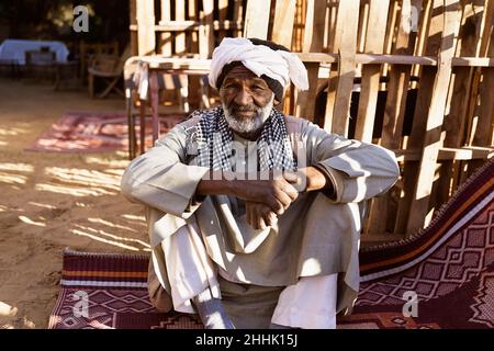 Homme nubien âgé dans une tenue ethnique assis sur un tapis dans la cour et regardant la caméra au soleil Banque D'Images