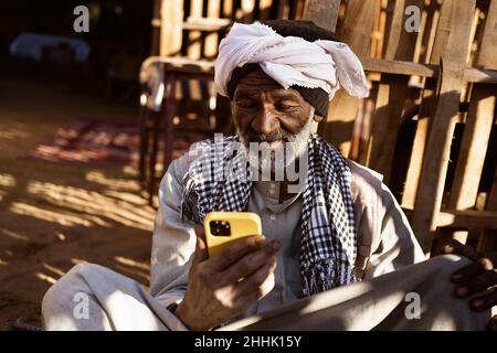 Homme nubien âgé en vêtements traditionnels et en turban assis dans la cour et utilisant le téléphone portable le jour ensoleillé Banque D'Images