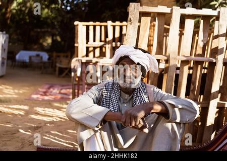 Homme nubien âgé dans une tenue ethnique assis sur un tapis dans la cour et regardant la caméra au soleil Banque D'Images