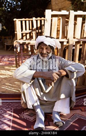 Homme nubien âgé dans une tenue ethnique assis sur un tapis dans la cour et regardant la caméra au soleil Banque D'Images