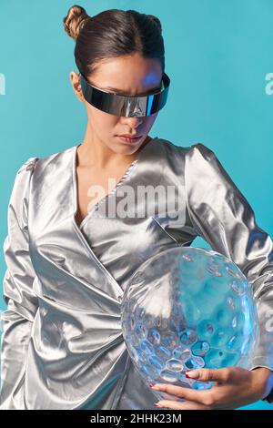 Jeune femme ethnique sérieuse avec des cheveux foncés dans une tenue tendance et lunettes VR futuriste boule de verre tout en explorant la réalité virtuelle contre le backgrou bleu Banque D'Images