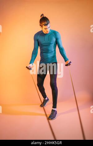 Pleine longueur de jeune tenue jeune hispanique athlète masculin avec des cheveux foncés dans les vêtements de sport exercice avec corde de résistance debout contre fond orange i Banque D'Images