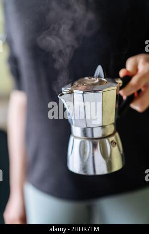 Attention douce de la récolte personne méconnaissable avec moka en métal de café chaud à la vapeur dans la main debout dans la cuisine à la maison Banque D'Images
