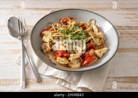 Spaghetti aux grains entiers avec tomates, champignons et lanières de poulet, garni de cresson, d'assiette grise et de couverts sur une table en bois rustique clair, sélectionnés Banque D'Images