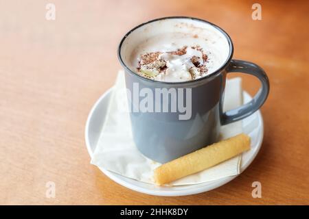 Chocolat chaud avec crème fouettée et saupoudrés de cacao servis avec un cookie dans une tasse bleue sur une table en bois, espace de copie, foyer sélectionné, étroit Banque D'Images