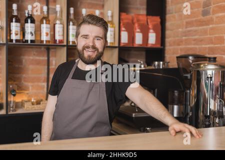 Barbu positif homme Barista dans un tablier près de l'appareil et des étagères de bouteilles en verre dans la maison de café et regardant l'appareil photo Banque D'Images