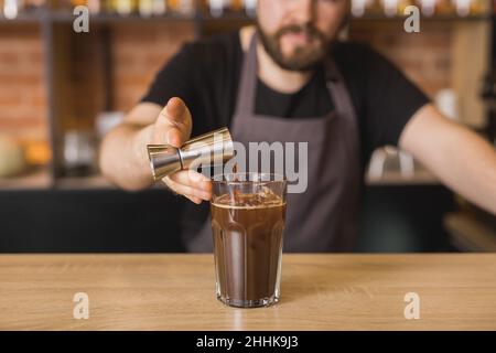 Croiser le barista barbu dans un tablier en versant le sirop du puzzle en métal dans un verre de café frais glacé au comptoir Banque D'Images