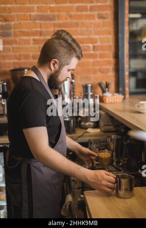 Vue latérale d'un Barista barbu en tablier prenant des tasses de boissons tout en travaillant au comptoir dans le café Banque D'Images