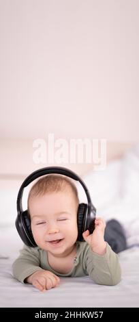 Bébé gai écoutant de la musique dans un casque couché sur un lit blanc, enfant de 3 mois petit dj, Happy enfance concept Banque D'Images