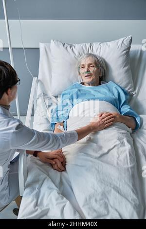 Vue en grand angle du médecin tenant les mains d'une femme âgée et la soutenant pendant la maladie à l'hôpital Banque D'Images