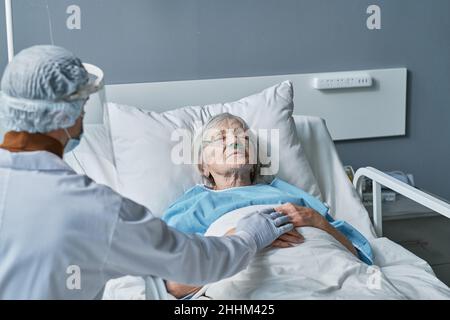 Médecin en port de protection s'occupant de la femme sénior malade pendant qu'elle dormait sur le lit à l'hôpital Banque D'Images