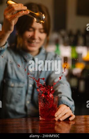 Femme barman ajoutant des glaçons dans le verre avec un cocktail alcoolisé à l'ancienne tout en se tenant au comptoir du bar pendant le travail Banque D'Images