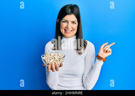 Jeune femme hispanique tenant des graines de citrouille bol souriant heureux pointant avec la main et le doigt sur le côté Banque D'Images