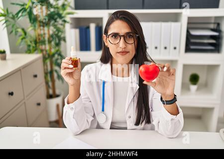 Jeune femme hispanique médecin tenant le coeur et l'huile de cbd détendue avec une expression sérieuse sur le visage. Simple et naturel regardant la caméra. Banque D'Images