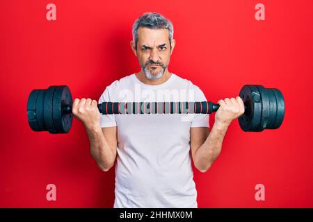 Beau homme d'âge moyen avec des cheveux gris portant des vêtements de sport en utilisant des haltères sceptique et nerveux, frowning bouleversé à cause du problème. Personne négative. Banque D'Images