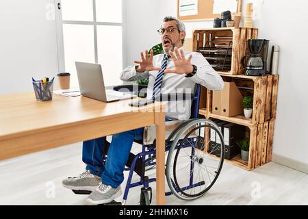 Homme hispanique d'âge moyen travaillant au bureau assis sur un fauteuil roulant peur et terrifié avec l'expression de peur arrêter le geste avec les mains, criant en sho Banque D'Images